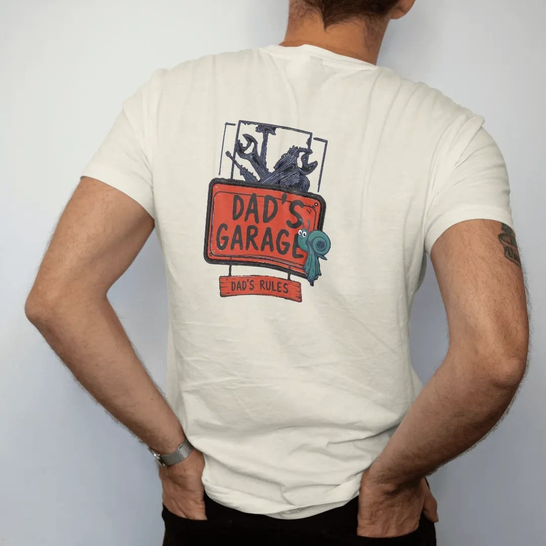 Man wearing a Ivory tee, seen from the back: a vintage garage sign with crossed wrenches over a pegboard and a hanging plank, Dad's Garage over Dad's Rules, in charcoal, rally red, navy and teal.