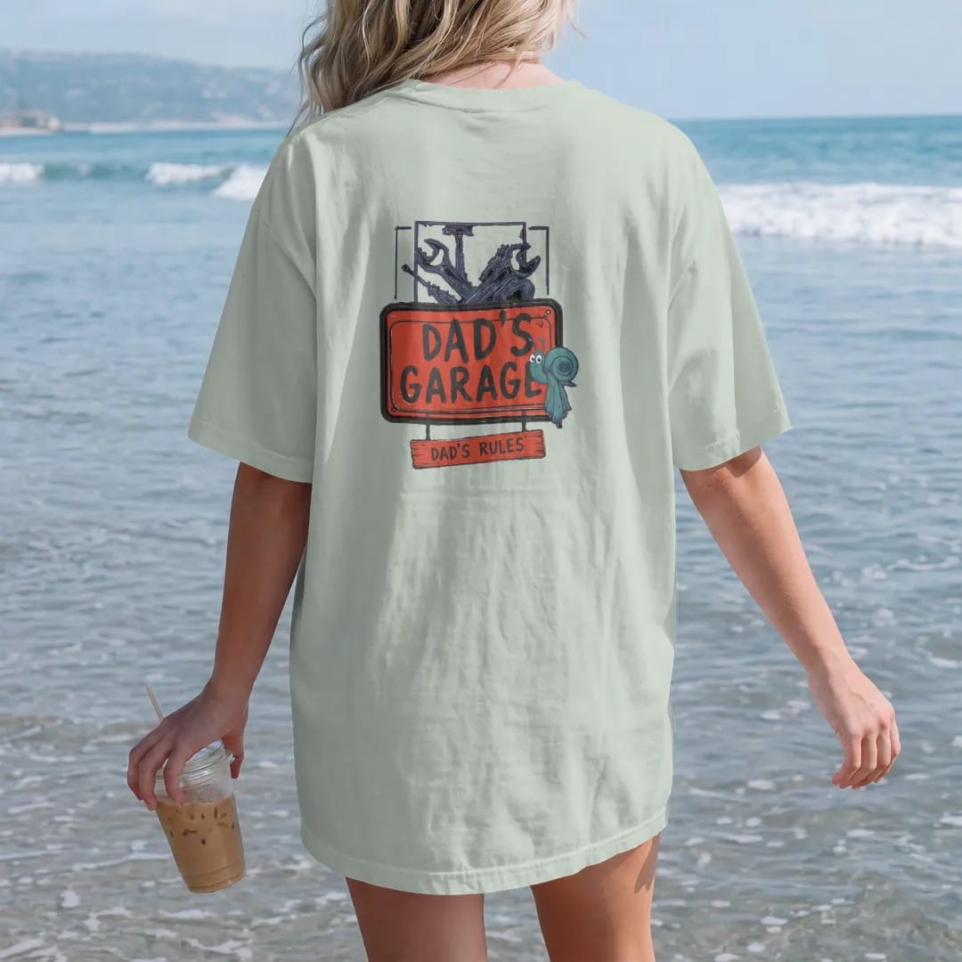 Woman on the beach in a Bay tee, back view: a vintage garage sign with crossed wrenches over a pegboard and a hanging plank, Dad's Garage over Dad's Rules, in charcoal, rally red, navy and teal.