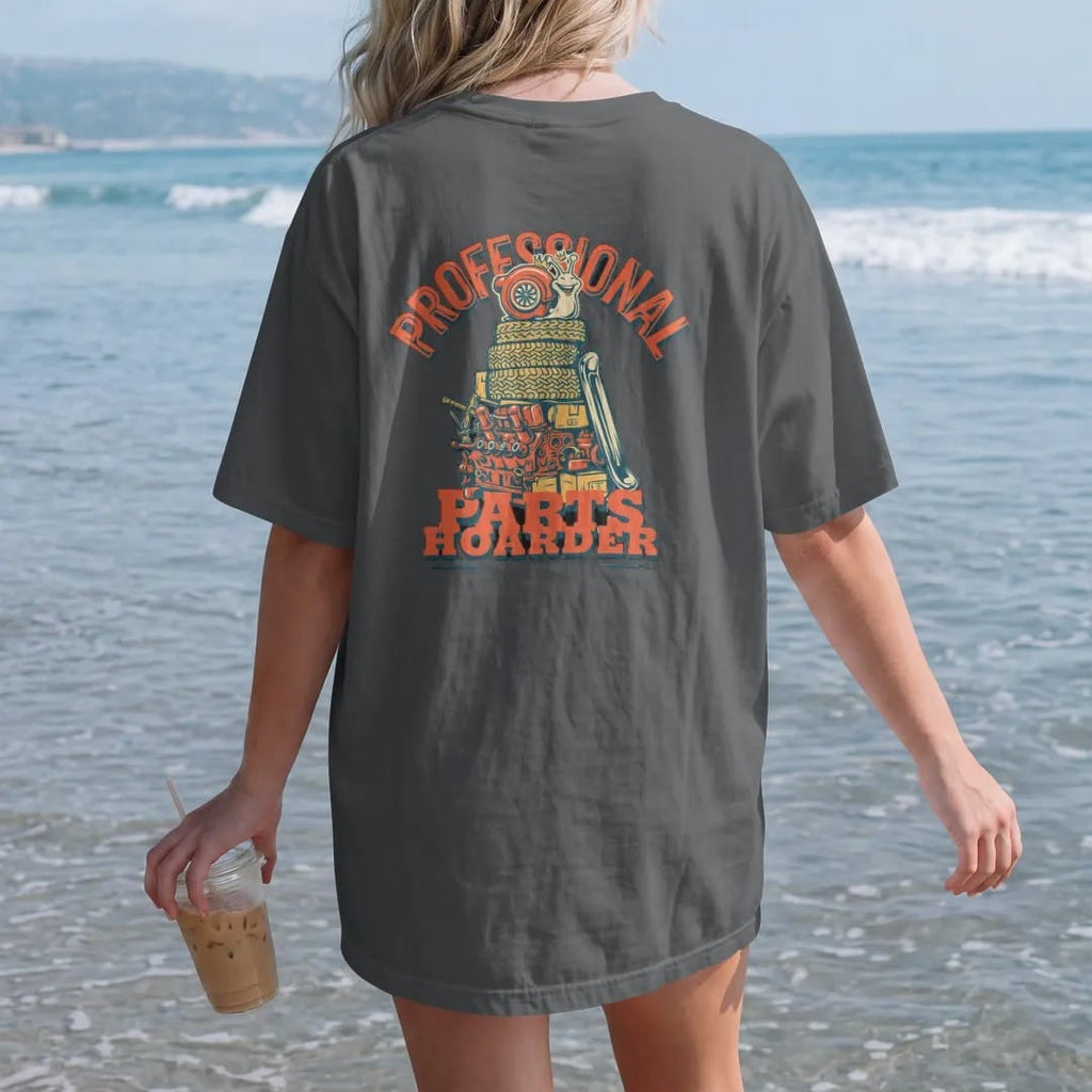 Woman on the beach in a Graphite tee, back view: the turbo snail mascot sitting on top of a towering pile of spare car parts and tires, Professional arched over Parts Hoarder, in cream, orange, yellow and teal.