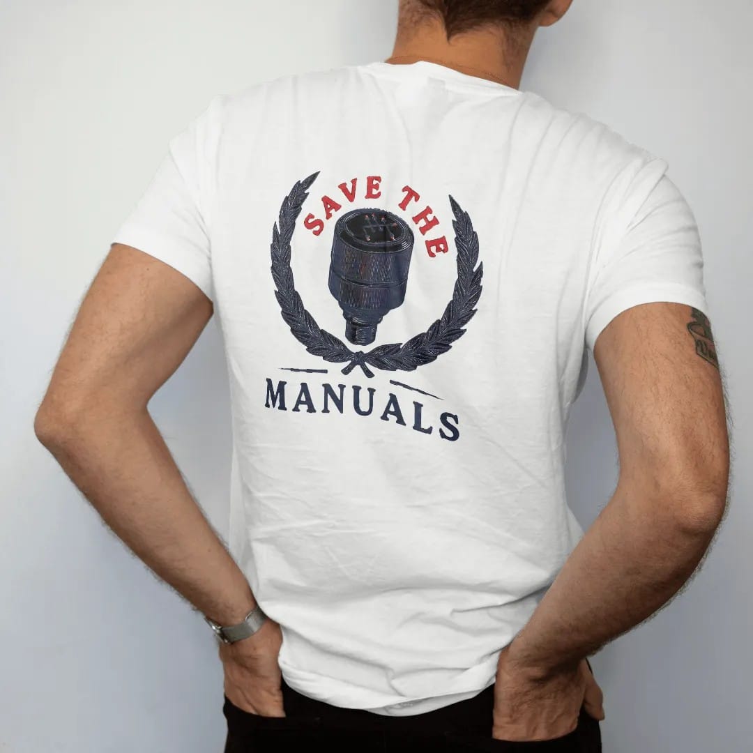 Man wearing a White tee, seen from the back: a vintage shift knob with a six-speed H-pattern inside a laurel wreath, Save The Manuals in engraved serif capitals, in navy, rally red and charcoal.
