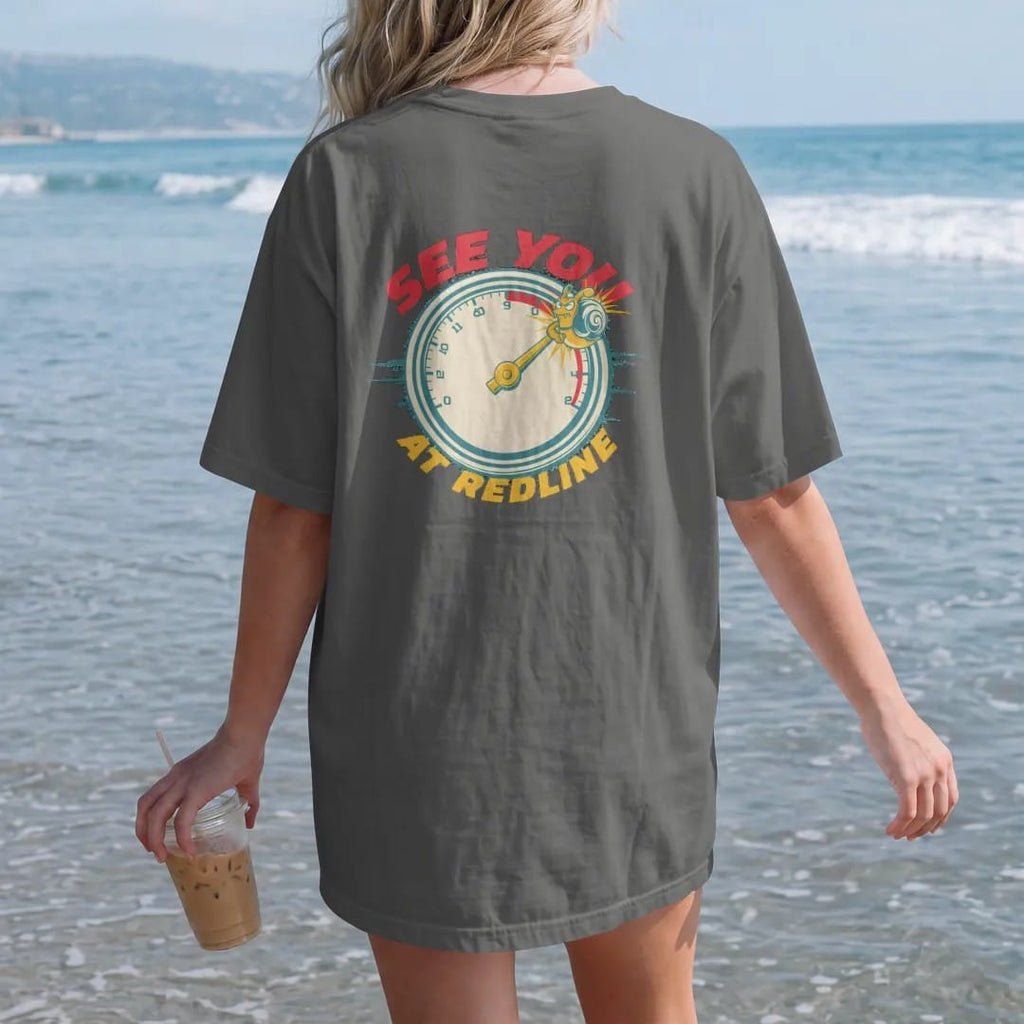 Woman on the beach in a Pepper tee, back view: a vintage tachometer with the needle in the red zone and the turbo snail clinging to the tip, See You At Redline lettering, in red, yellow, cream and teal.