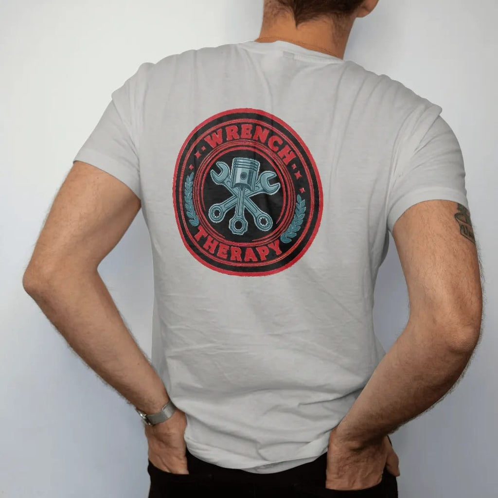 Man wearing a Granite tee, seen from the back: a round vintage badge with crossed wrenches over a piston and laurel leaves, Wrench arched over Therapy, in charcoal, rally red and teal.