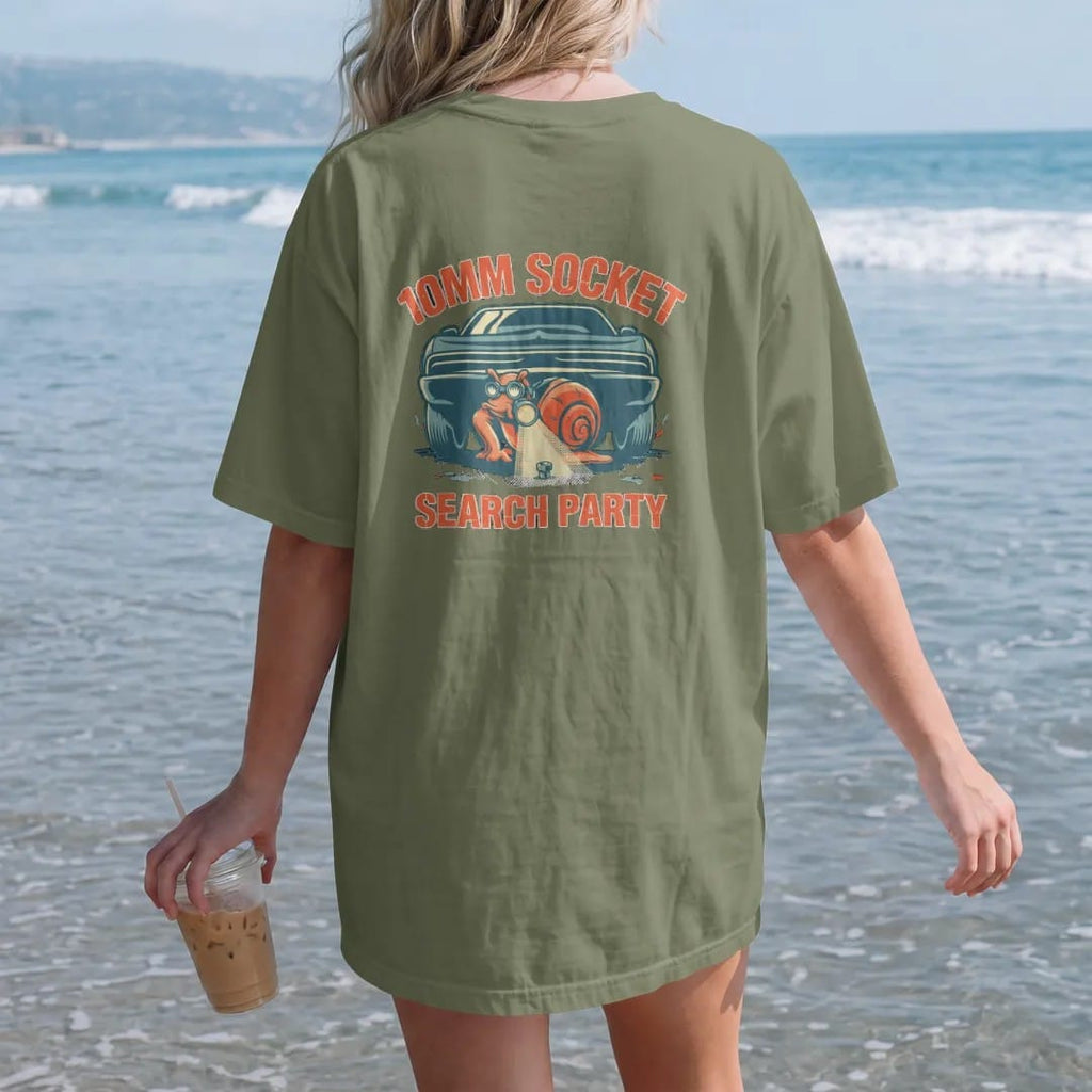 Woman on the beach in a Moss tee, back view: the turbo snail mascot with a headlamp and flashlight finding a lost socket under a generic car, 10mm Socket Search Party lettering, in cream, yellow, orange and teal.