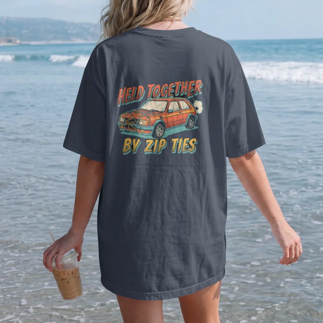 Woman on the beach in a Midnight tee, back view: a generic 90s hatchback with its bumper and hood held on by bright zip ties, Held Together By Zip Ties lettering, in cream, orange, yellow and teal.