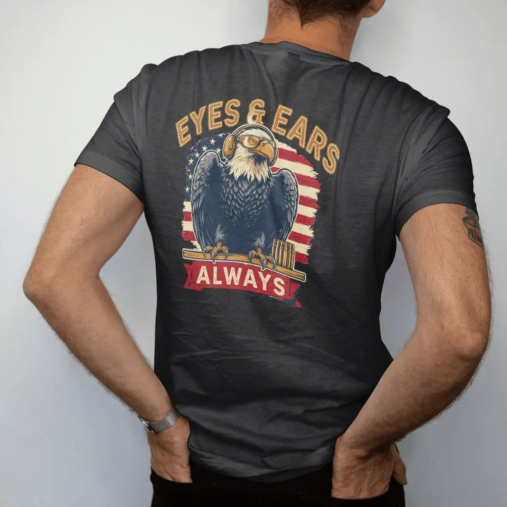 Man wearing a Black tee, seen from the back: a bald eagle in hearing protection and shooting glasses perched on a range bench with brass cartridges, Eyes & Ears arched over an Always banner, in brass gold, cream, red and faded blue.