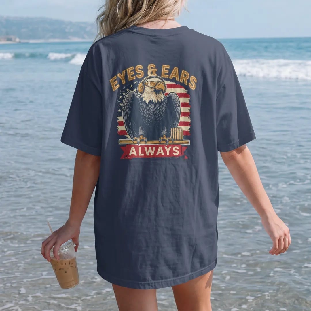 Woman on the beach in a Navy tee, back view: a bald eagle in hearing protection and shooting glasses perched on a range bench with brass cartridges, Eyes & Ears arched over an Always banner, in brass gold, cream, red and faded blue.