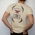 Man wearing a Sandstone tee, seen from the back: a vintage ammo-box label layout with five upright brass cartridges under a small eagle crest, Brass & Powder Co. over Fine Cartridges, in navy, oxblood and olive drab.