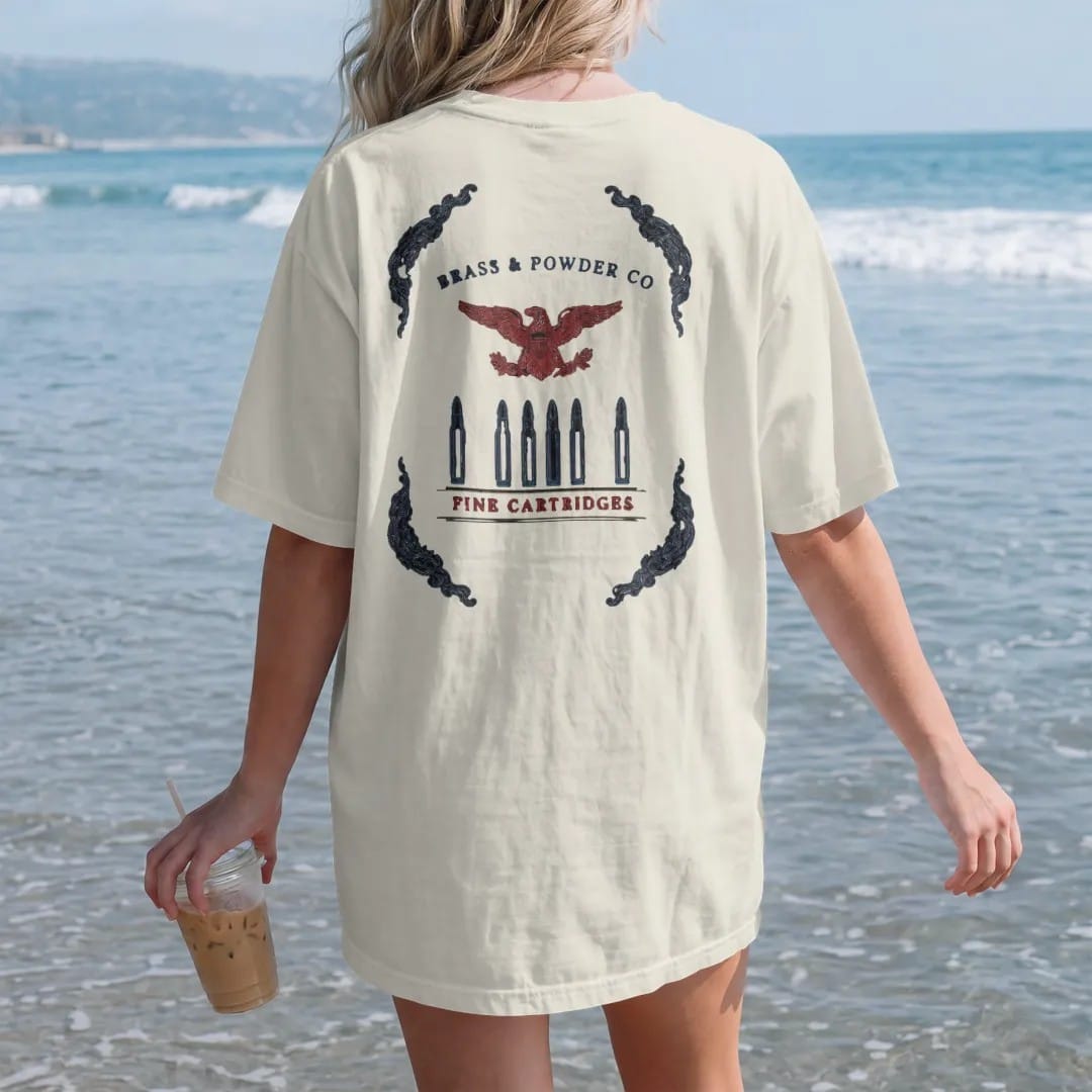 Woman on the beach in a Ivory tee, back view: a vintage ammo-box label layout with five upright brass cartridges under a small eagle crest, Brass & Powder Co. over Fine Cartridges, in navy, oxblood and olive drab.