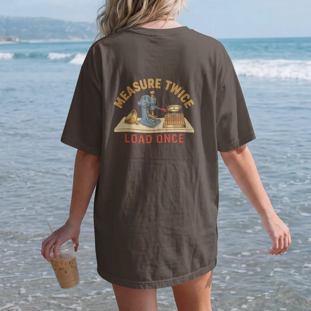 Woman on the beach in a Espresso tee, back view: a generic reloading press on a wooden bench with a powder scale, funnel and rows of brass cases, Measure Twice Load Once lettering, in brass gold, cream, blue and red.