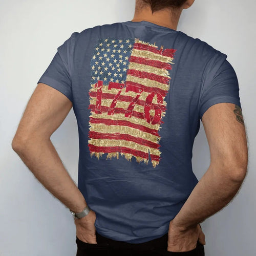 Man wearing a Navy tee, seen from the back: a distressed American flag with stripes made of brass cartridges and 1776 in big stencil numerals, in brass gold, cream, red and faded blue.