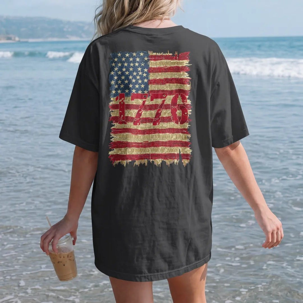 Woman on the beach in a Black tee, back view: a distressed American flag with stripes made of brass cartridges and 1776 in big stencil numerals, in brass gold, cream, red and faded blue.