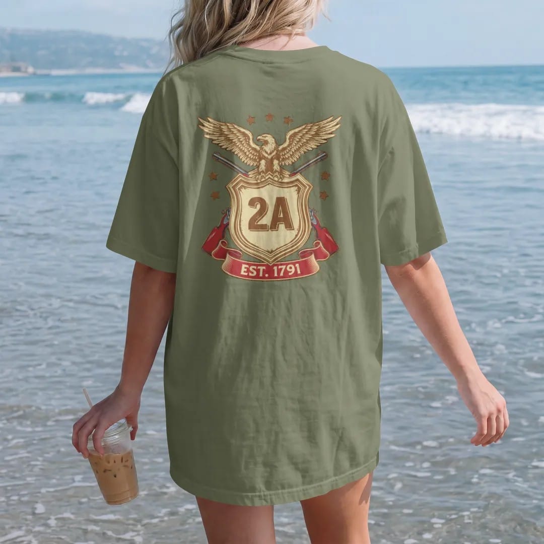 Woman on the beach in a Moss tee, back view: a heraldic shield with 2A at the center over two crossed generic rifles and a spread-winged eagle, Est. 1791 on a ribbon, in brass gold, cream, red and blue.