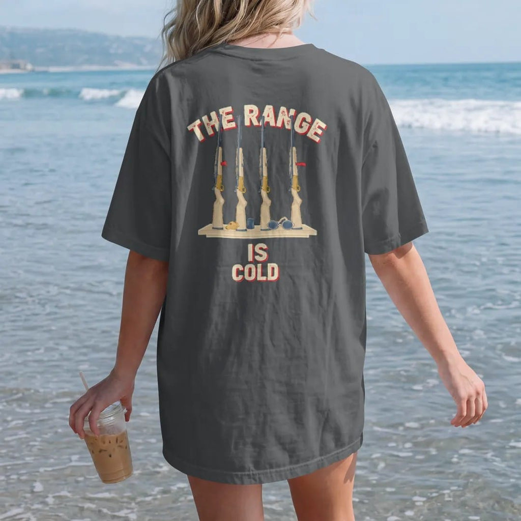 Woman on the beach in a Graphite tee, back view: a rack of generic rifles with open actions and chamber flags beside a whistle and safety glasses, The Range Is Cold lettering, in cream, red, brass gold and blue.