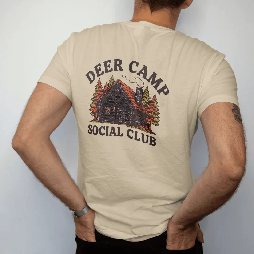 Man wearing a Sandstone tee, seen from the back: a rustic log cabin in autumn woods with chimney smoke, Deer Camp arched above and Social Club below.