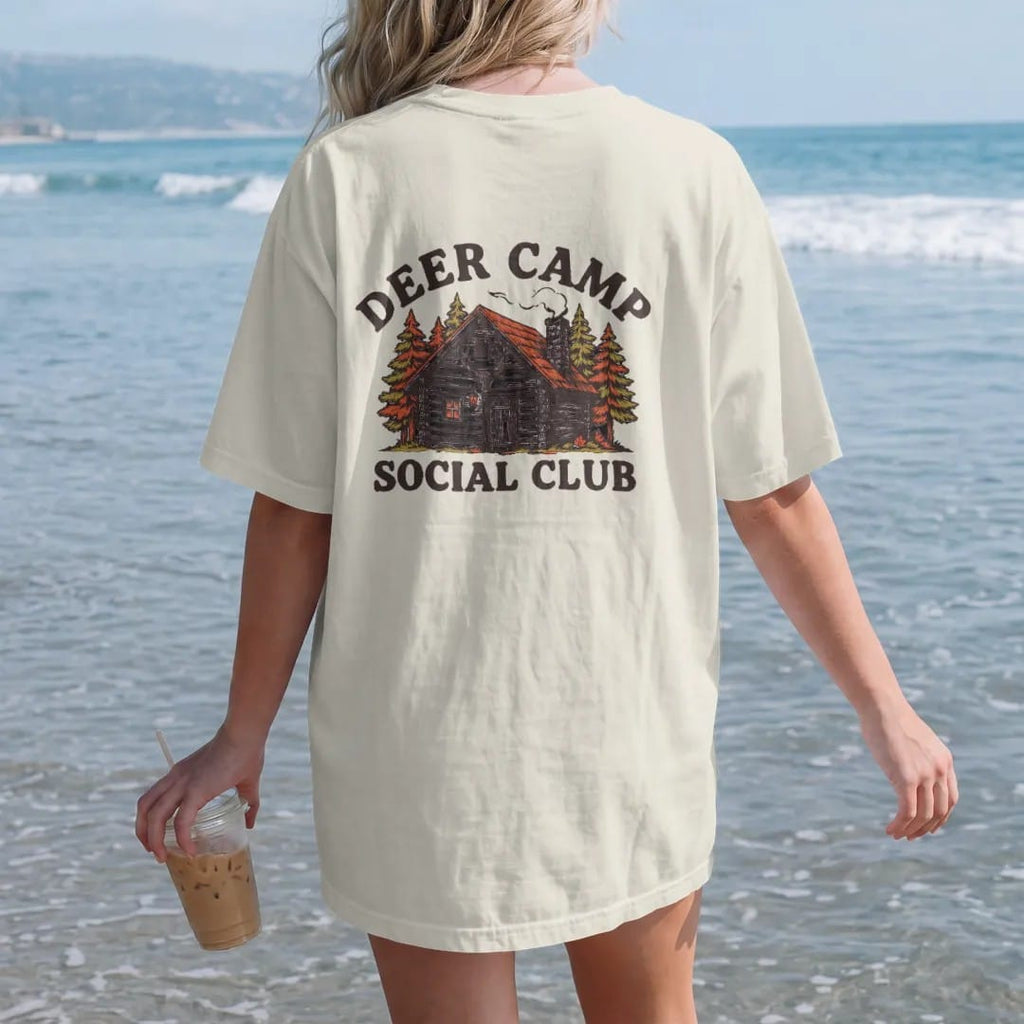 Woman on the beach in a Ivory tee, back view: a rustic log cabin in autumn woods with chimney smoke, Deer Camp arched above and Social Club below.