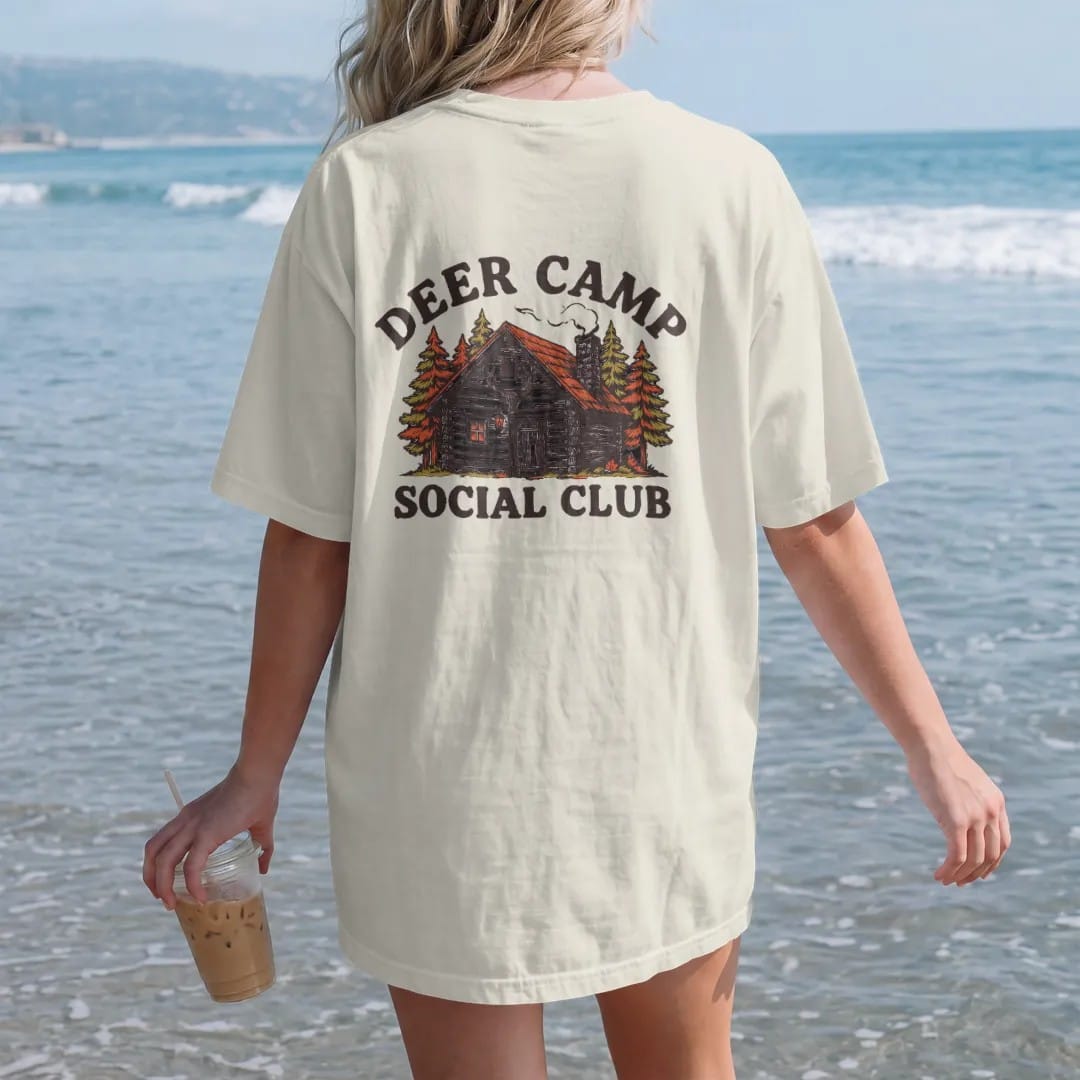 Woman on the beach in a Ivory tee, back view: a rustic log cabin in autumn woods with chimney smoke, Deer Camp arched above and Social Club below.