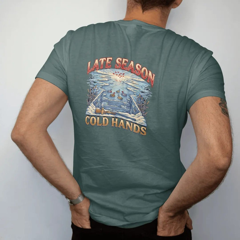 Man wearing a Blue Spruce tee, seen from the back: a snowy duck blind at dawn with mallard decoys, a steaming thermos and a generic shotgun resting on the rail, Late Season Cold Hands lettering, in cream, faded blue, brass gold and red.