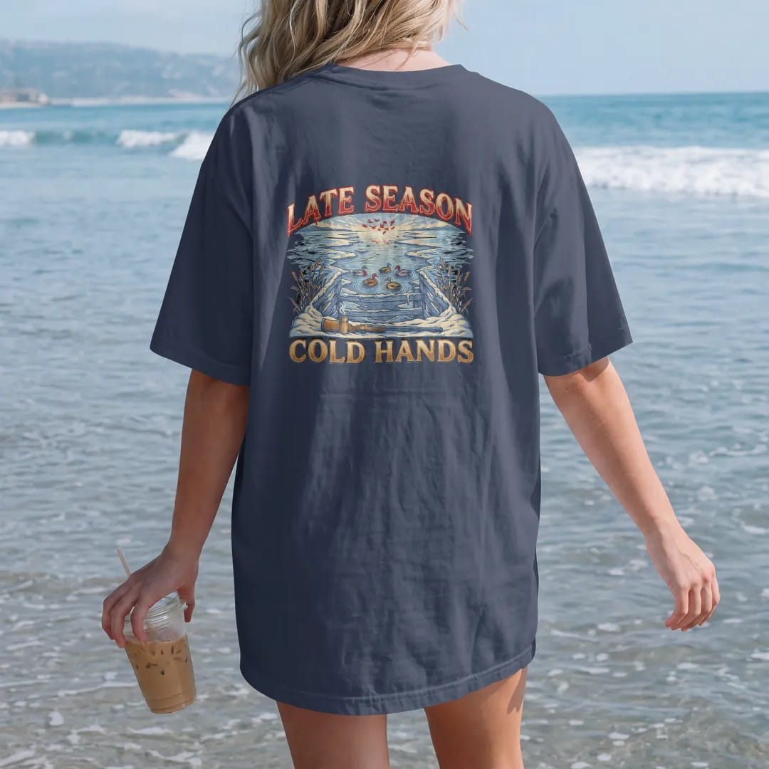 Woman on the beach in a Navy tee, back view: a snowy duck blind at dawn with mallard decoys, a steaming thermos and a generic shotgun resting on the rail, Late Season Cold Hands lettering, in cream, faded blue, brass gold and red.