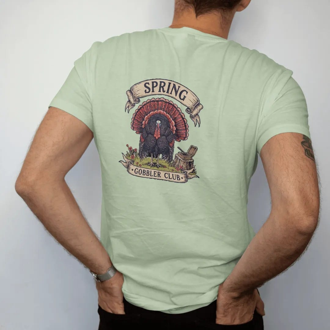 Man wearing a Light Green tee, seen from the back: a strutting wild turkey in a spring field with wildflowers and a box call, Spring Gobbler Club lettering, in olive drab, oxblood, navy and charcoal.