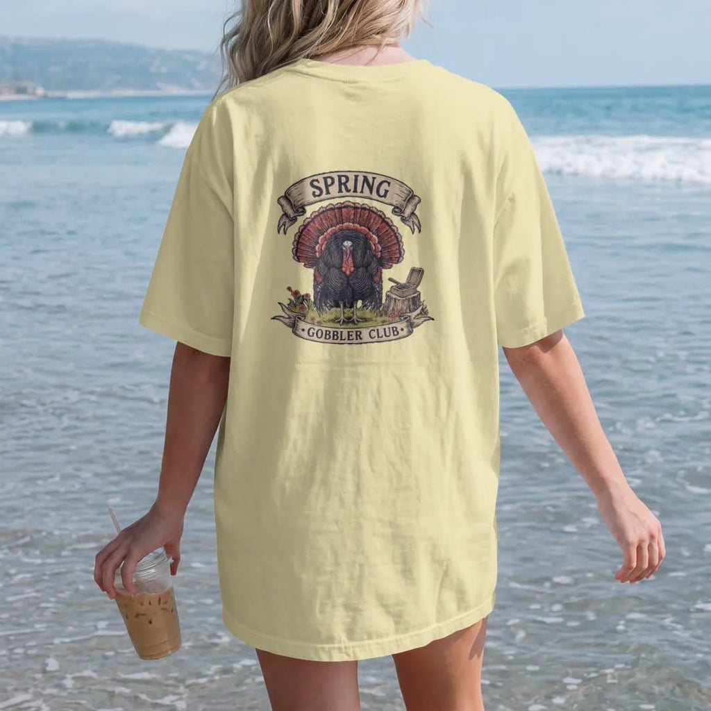 Woman on the beach in a Butter tee, back view: a strutting wild turkey in a spring field with wildflowers and a box call, Spring Gobbler Club lettering, in olive drab, oxblood, navy and charcoal.