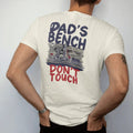 Man wearing a Ivory tee, seen from the back: a tidy reloading bench with a press, scale and brass, with Dad's Bench in big hand-painted letters above and Don't Touch below.