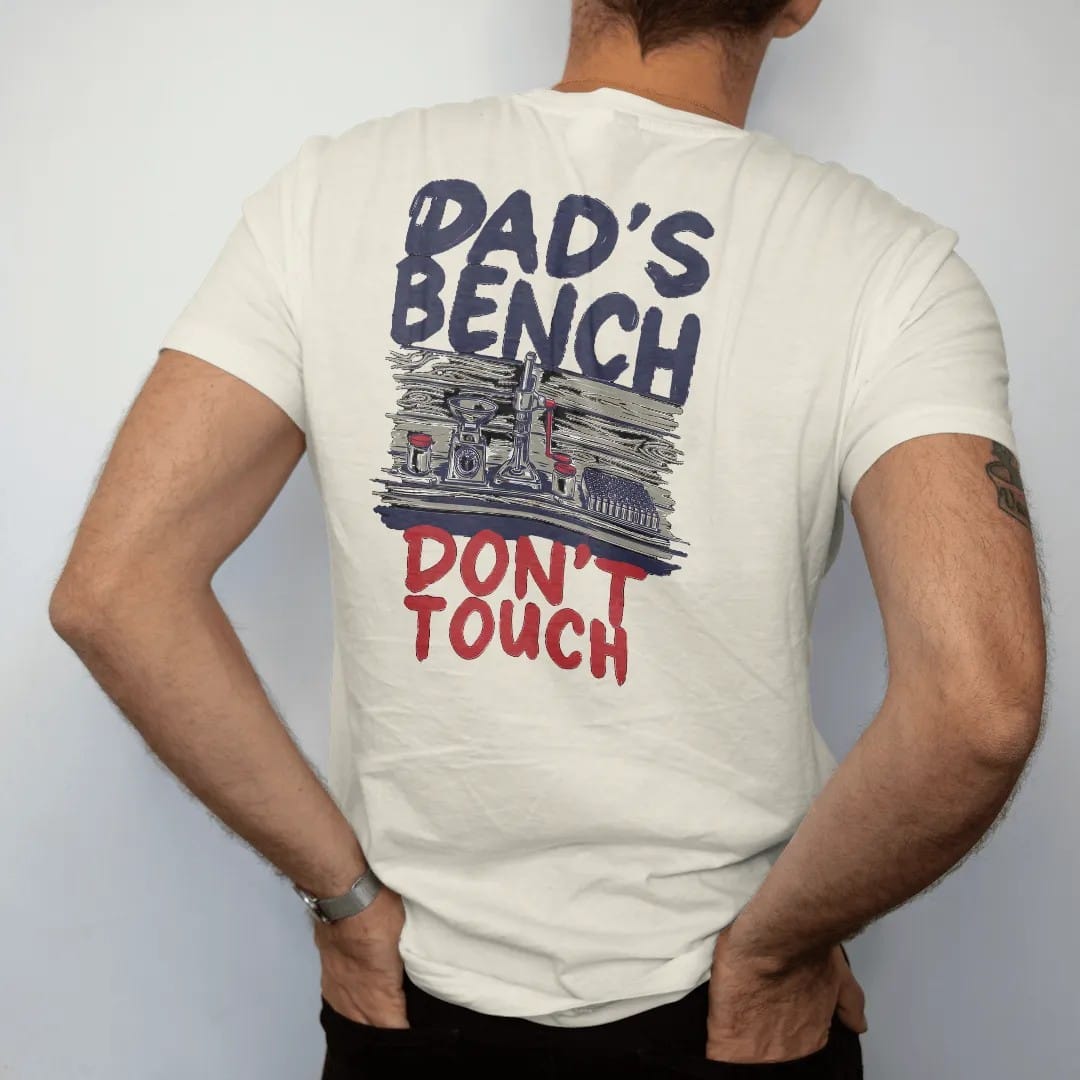 Man wearing a Ivory tee, seen from the back: a tidy reloading bench with a press, scale and brass, with Dad's Bench in big hand-painted letters above and Don't Touch below.