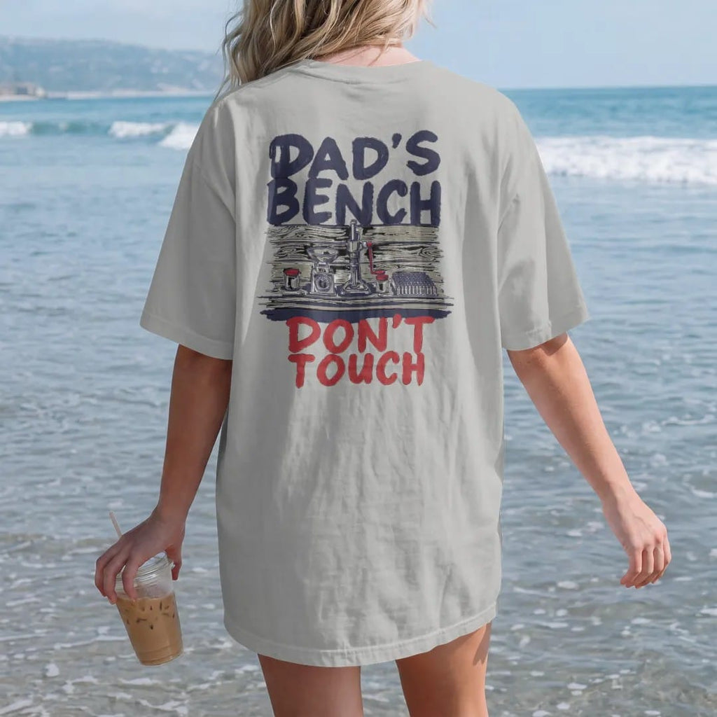 Woman on the beach in a Granite tee, back view: a tidy reloading bench with a press, scale and brass, with Dad's Bench in big hand-painted letters above and Don't Touch below.
