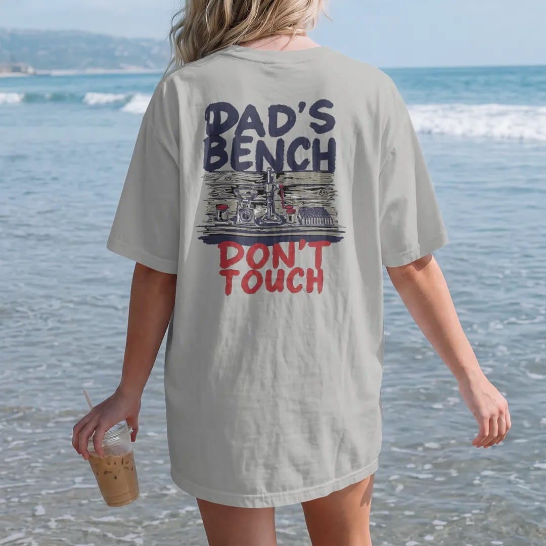 Woman on the beach in a Granite tee, back view: a tidy reloading bench with a press, scale and brass, with Dad's Bench in big hand-painted letters above and Don't Touch below.