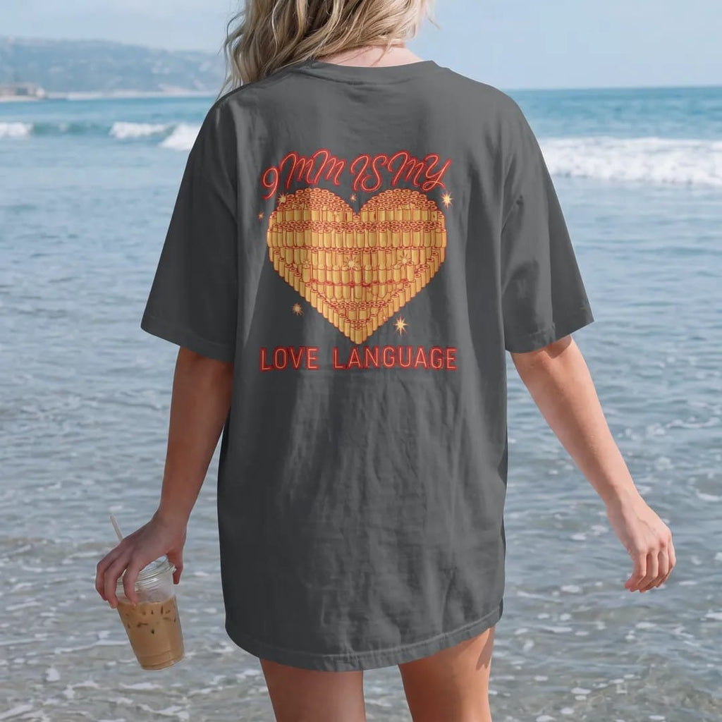 Woman on the beach in a Graphite tee, back view: a tattoo-flash heart made of stacked 9mm brass cartridges with sparkles, 9mm Is My Love Language lettering, in brass gold, red and cream.