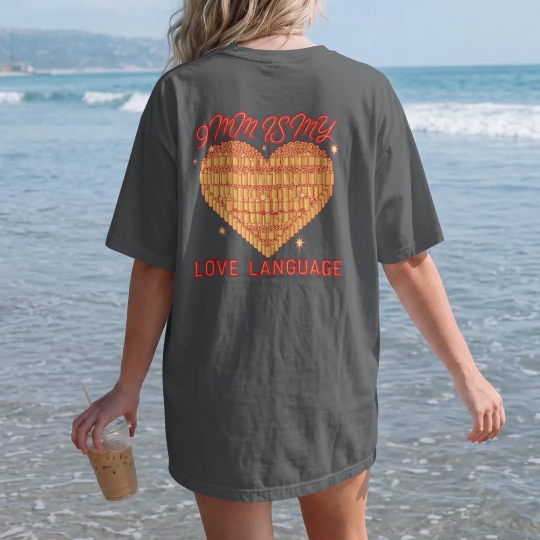 Woman on the beach in a Graphite tee, back view: a tattoo-flash heart made of stacked 9mm brass cartridges with sparkles, 9mm Is My Love Language lettering, in brass gold, red and cream.