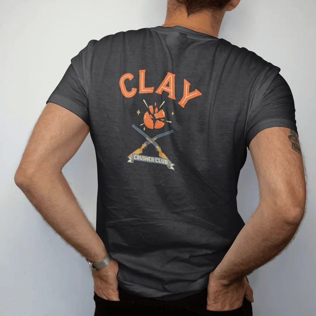 Man wearing a Black tee, seen from the back: an orange clay target shattering mid-air above two crossed generic shotguns, Clay Crusher Club lettering, in safety orange, cream, brass gold and blue.
