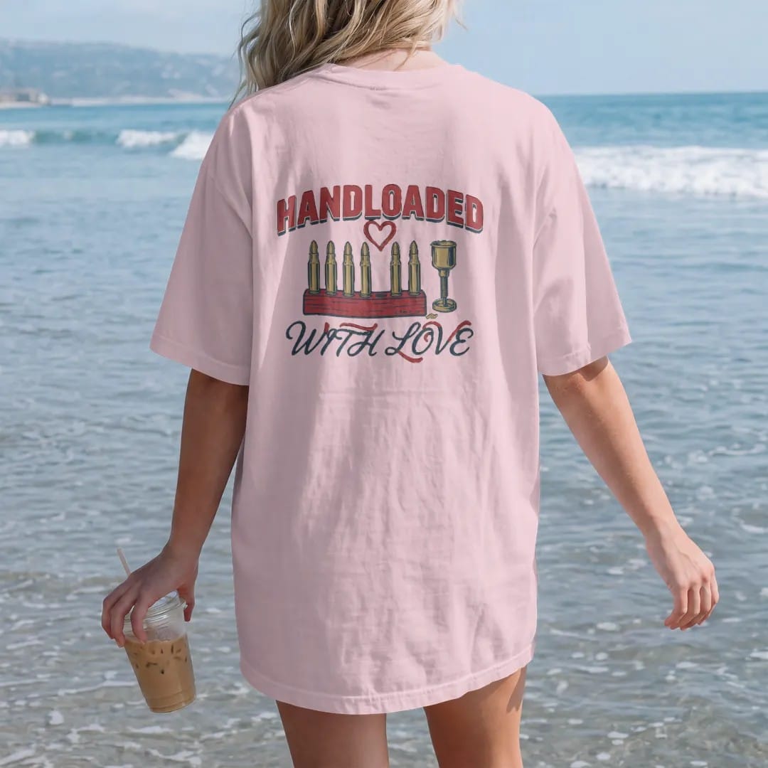 Woman on the beach in a Blossom tee, back view: a row of handloaded brass cartridges in a wooden loading block with a heart and a powder measure, Handloaded With Love lettering, in oxblood, navy and olive drab.