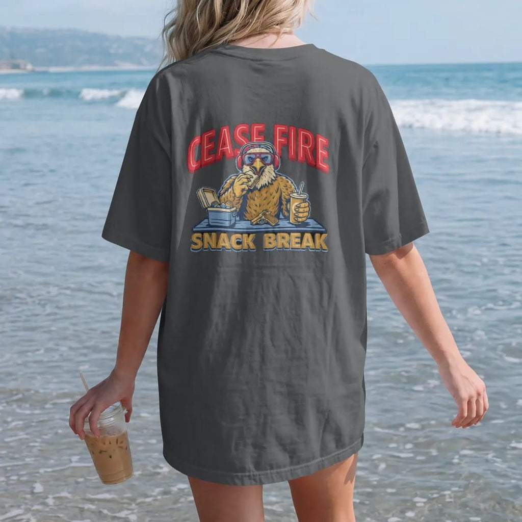 Woman on the beach in a Graphite tee, back view: the bald eagle mascot in hearing protection eating a sandwich behind a range bench and cooler with a generic pistol locked open, Cease Fire Snack Break lettering, in brass gold, cream, red and blue.