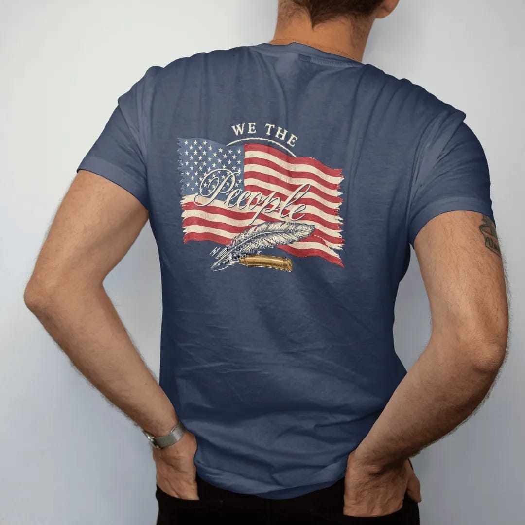 Man wearing a Navy tee, seen from the back: a distressed American flag with We The People in 18th-century script over a crossed quill and brass cartridge, in cream, red, faded blue and brass gold.
