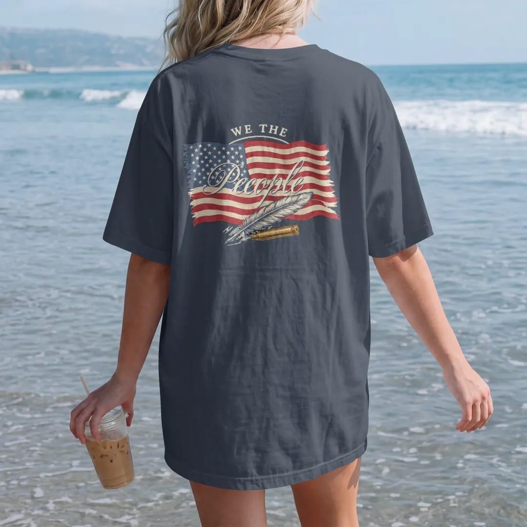Woman on the beach in a Midnight tee, back view: a distressed American flag with We The People in 18th-century script over a crossed quill and brass cartridge, in cream, red, faded blue and brass gold.