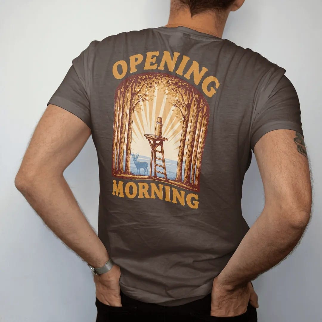 Man wearing a Espresso tee, seen from the back: a tree stand with a thermos in a hardwood forest at sunrise with a distant whitetail buck in the mist, Opening Morning lettering, in brass gold, cream, burnt orange and blue.