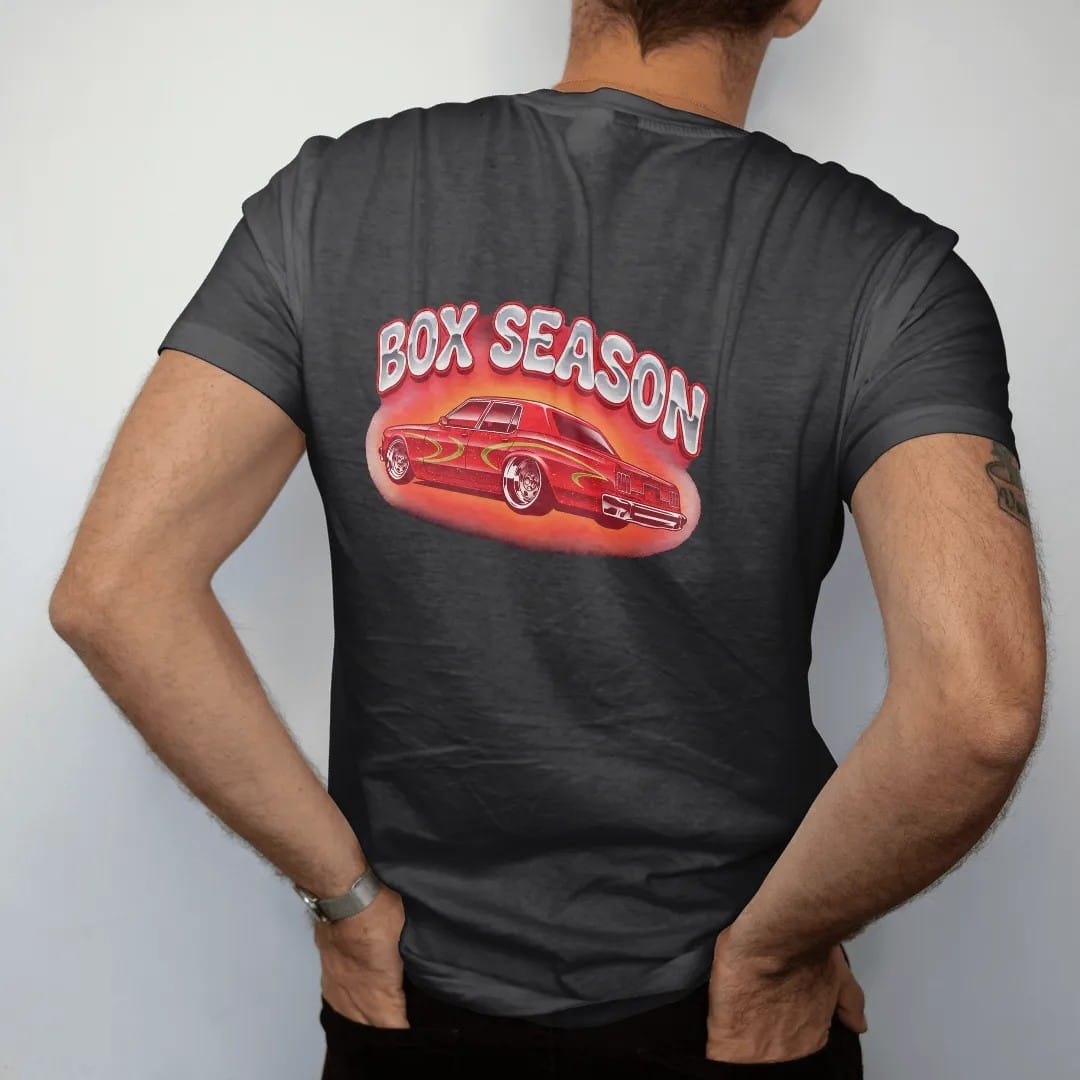 Man wearing a Black tee, seen from the back: a candy red boxy 80s sedan sitting high on big chrome rims under Box Season in chrome bubble letters, in red, silver, orange and lime.