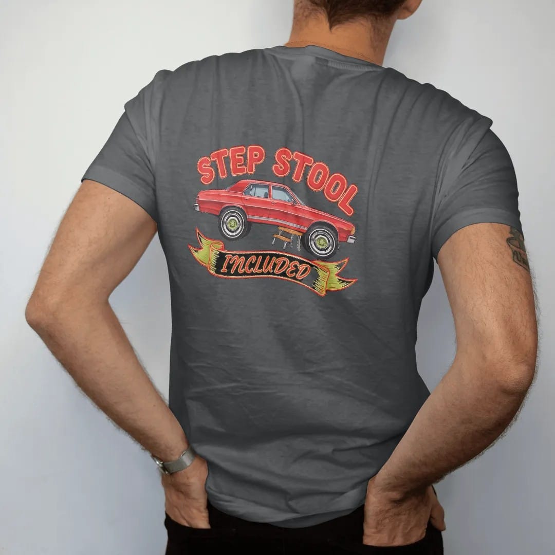 Man wearing a Graphite tee, seen from the back: a boxy 80s sedan sitting sky high on giant chrome rims with a little step stool by the door, under Step Stool and over a banner reading Included.
