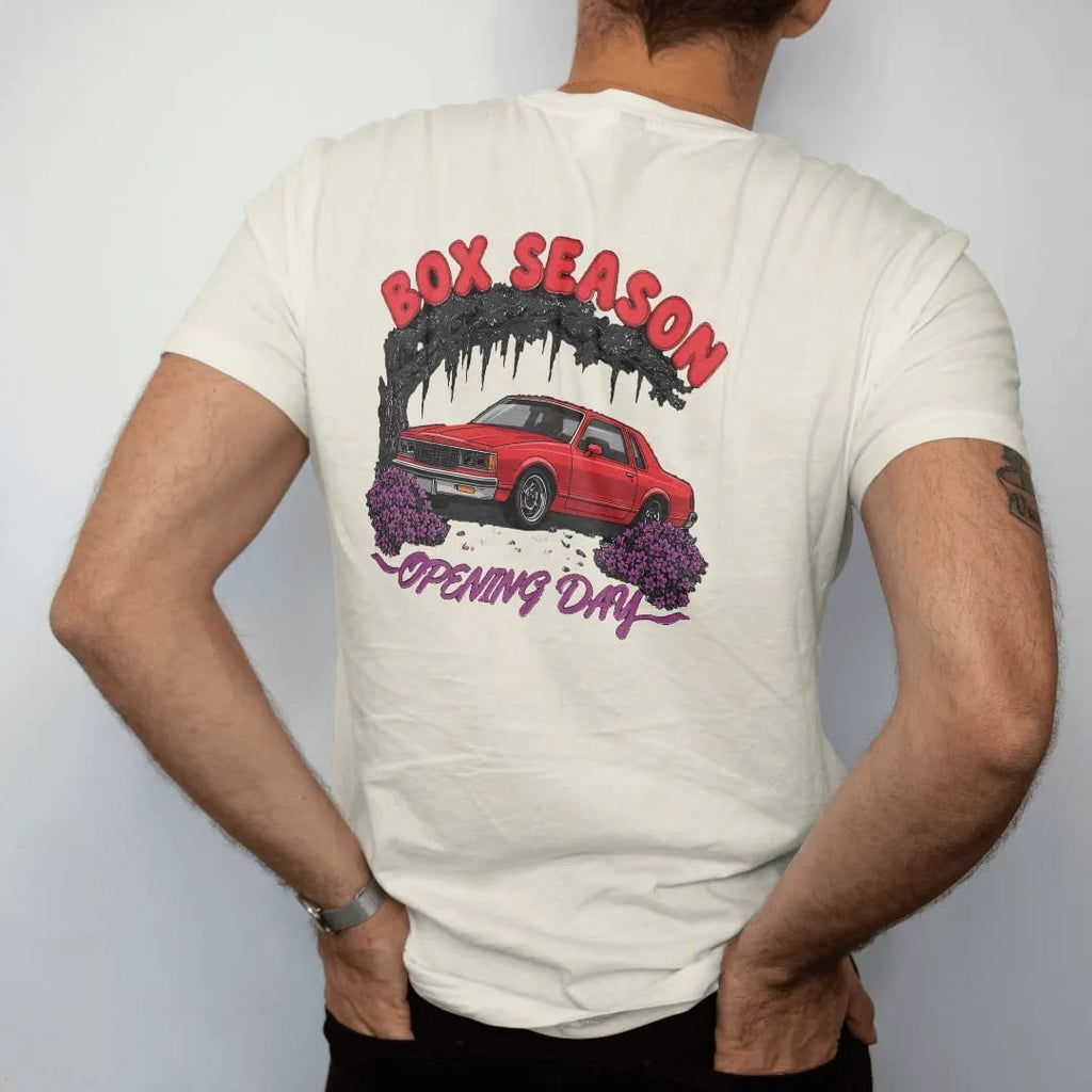 Man wearing a Ivory tee, seen from the back: a boxy 80s sedan sitting high among blooming azaleas and mossy live oaks, under Box Season and over Opening Day, in deep red, purple and gunmetal.