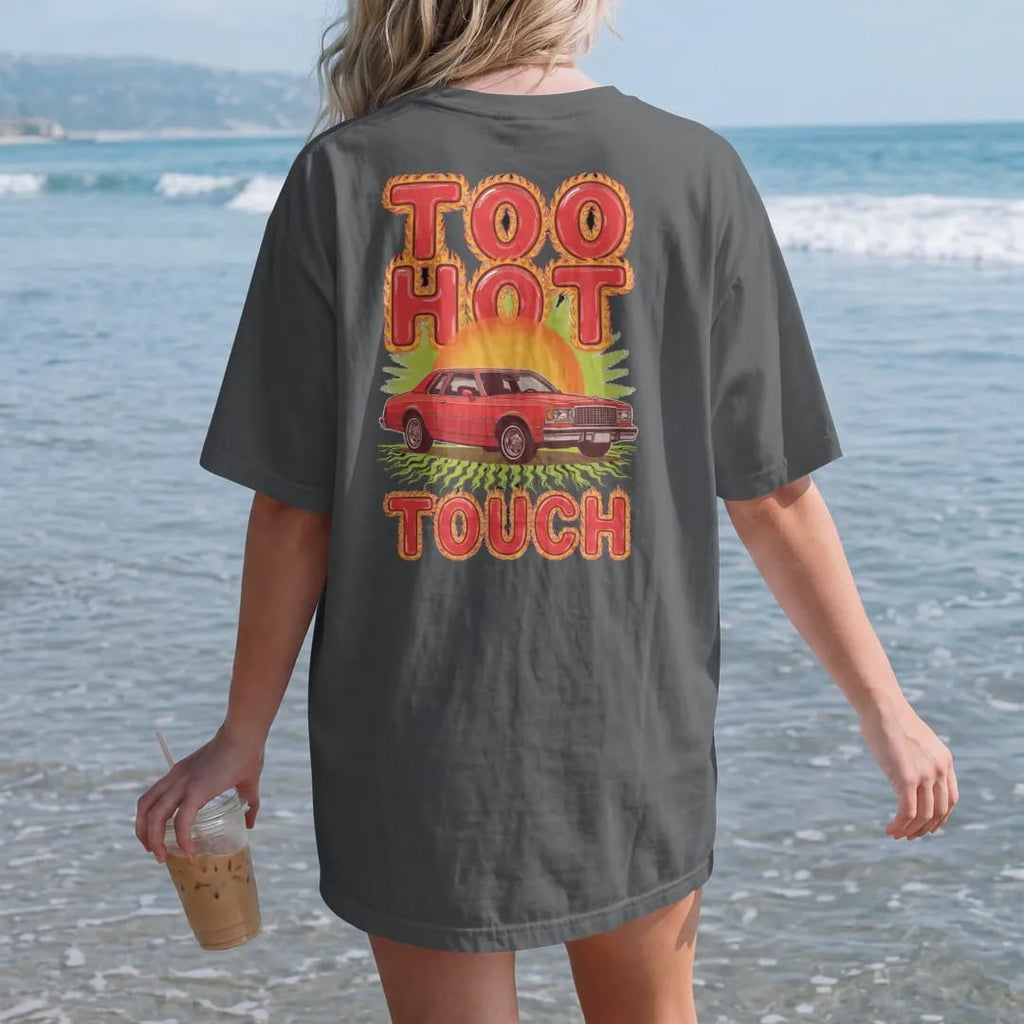 Woman on the beach in a Graphite tee, back view: a boxy 80s sedan on big chrome rims on shimmering hot asphalt under a blazing sun, with Too Hot over To Touch in flame-edged letters.