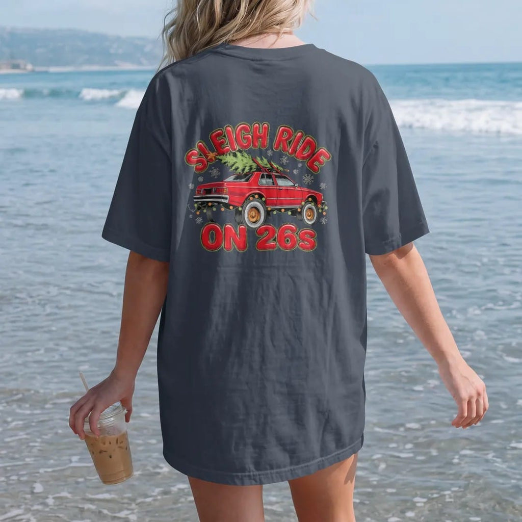 Woman on the beach in a Midnight tee, back view: a boxy 80s sedan on giant chrome rims with a Christmas tree on the roof and string lights in falling snow, under Sleigh Ride and over On 26s.