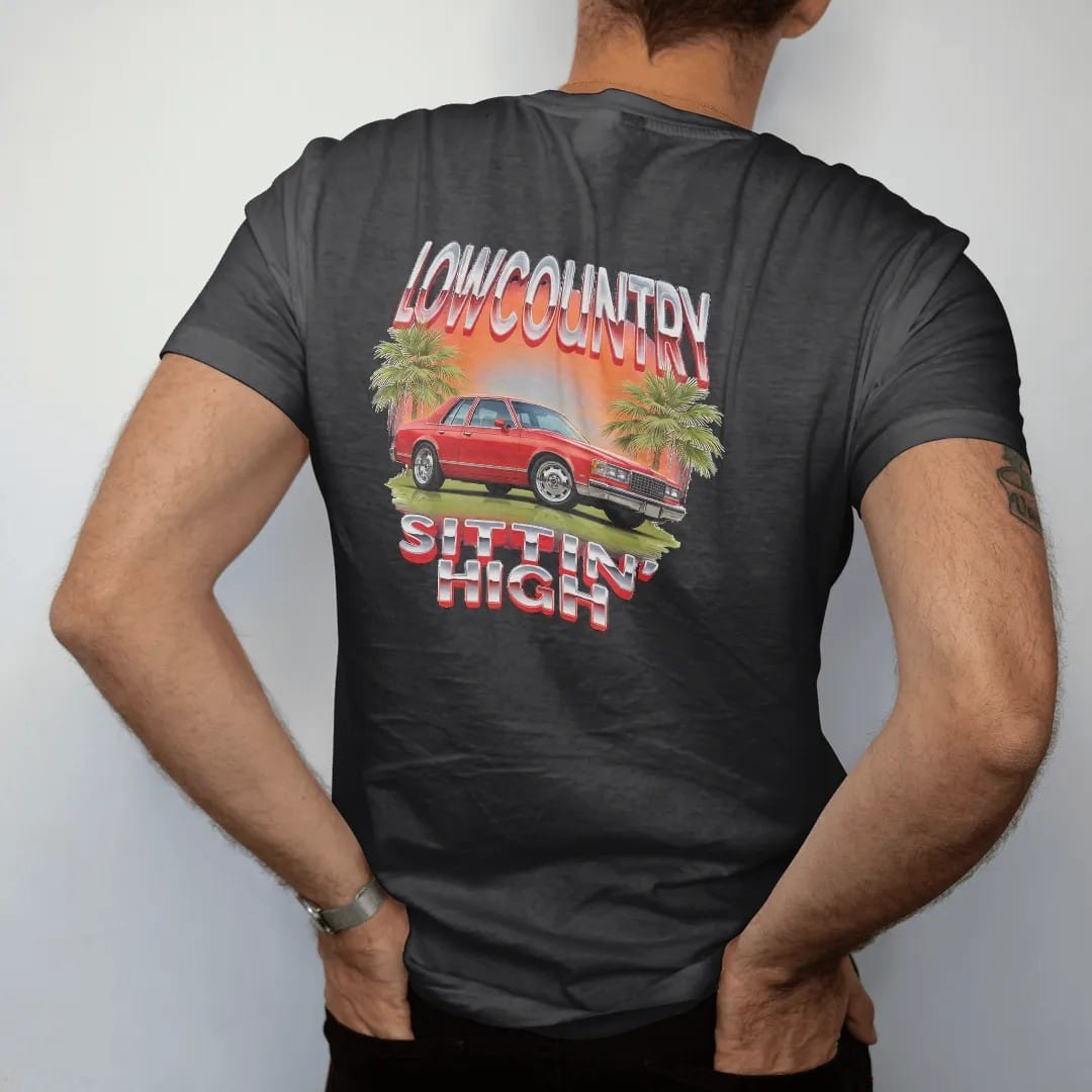 Man wearing a Black tee, seen from the back: a 90s airbrush style scene of a boxy 80s sedan sitting high on a marsh road at sunset with palmettos, under Lowcountry and over Sittin' High.