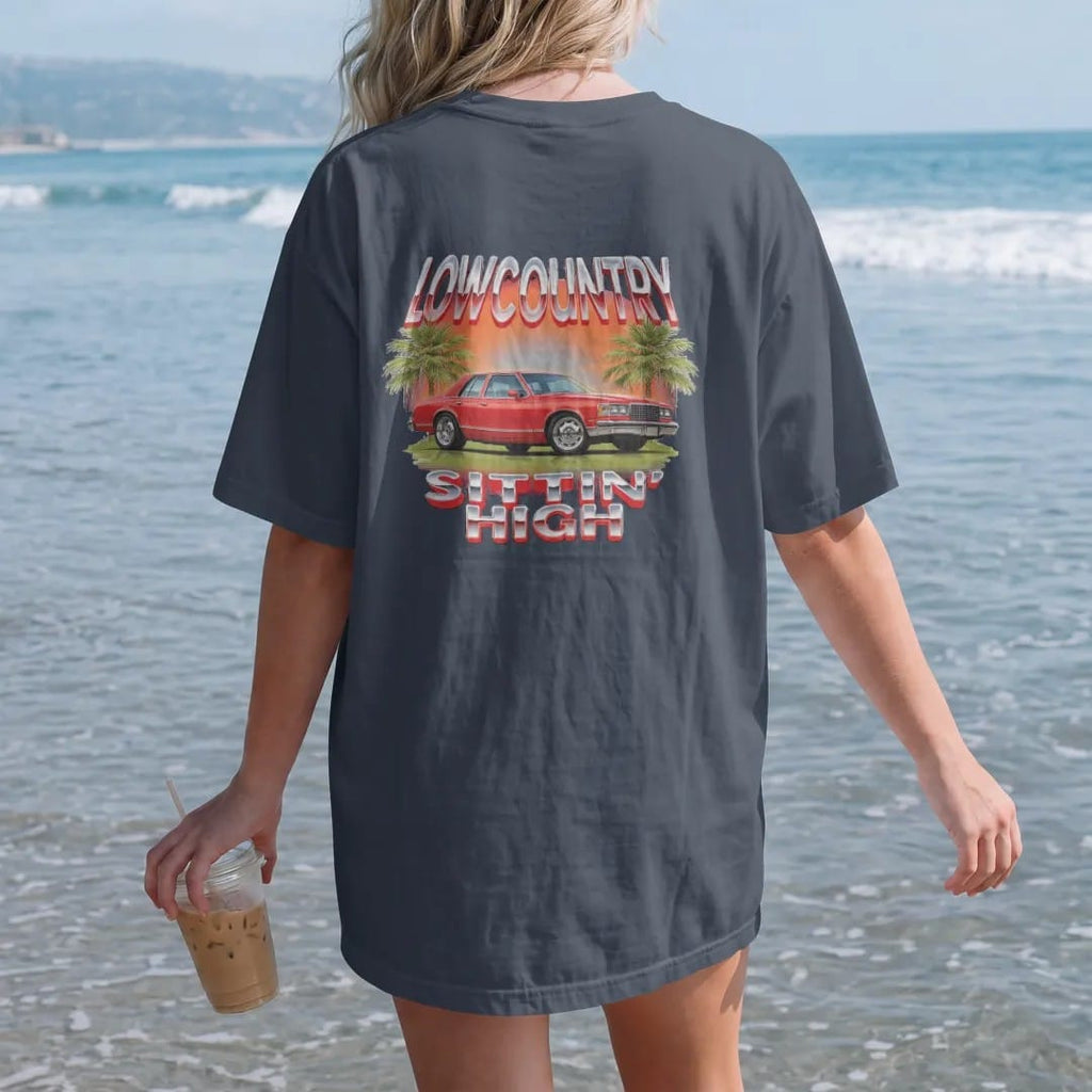 Woman on the beach in a Midnight tee, back view: a 90s airbrush style scene of a boxy 80s sedan sitting high on a marsh road at sunset with palmettos, under Lowcountry and over Sittin' High.