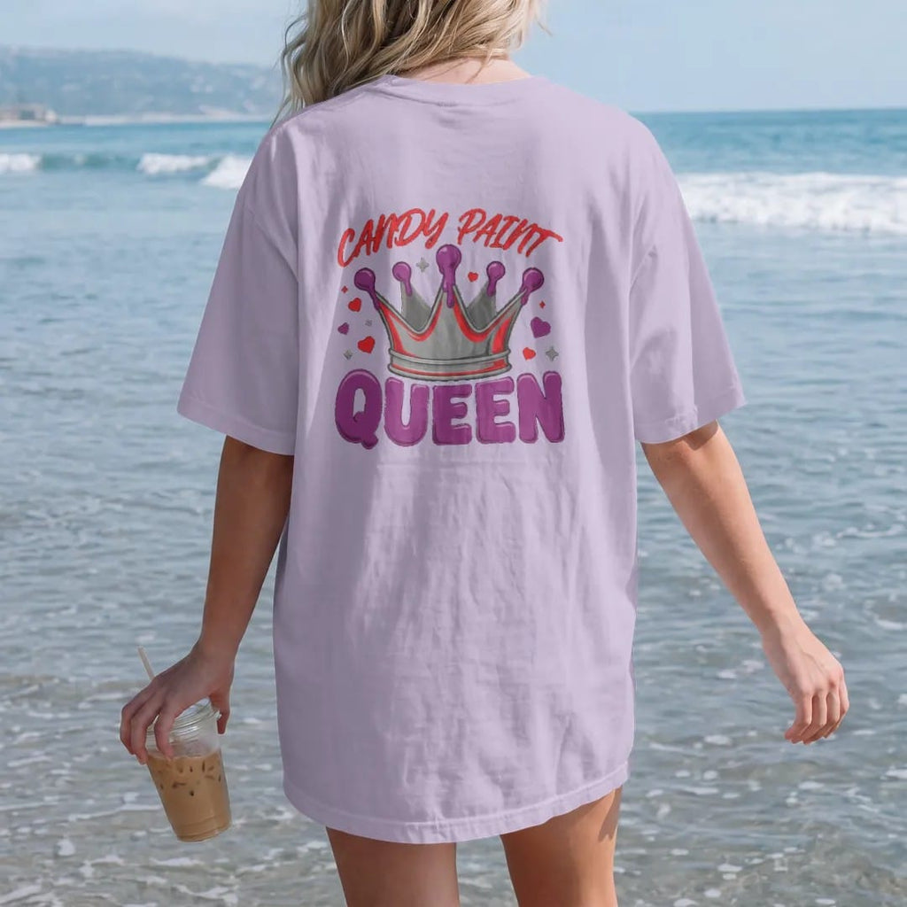 Woman on the beach in a Orchid tee, back view: a sparkling crown dripping candy paint on top of a chrome rim with hearts, under Candy Paint and over Queen, in deep red, purple and gunmetal.