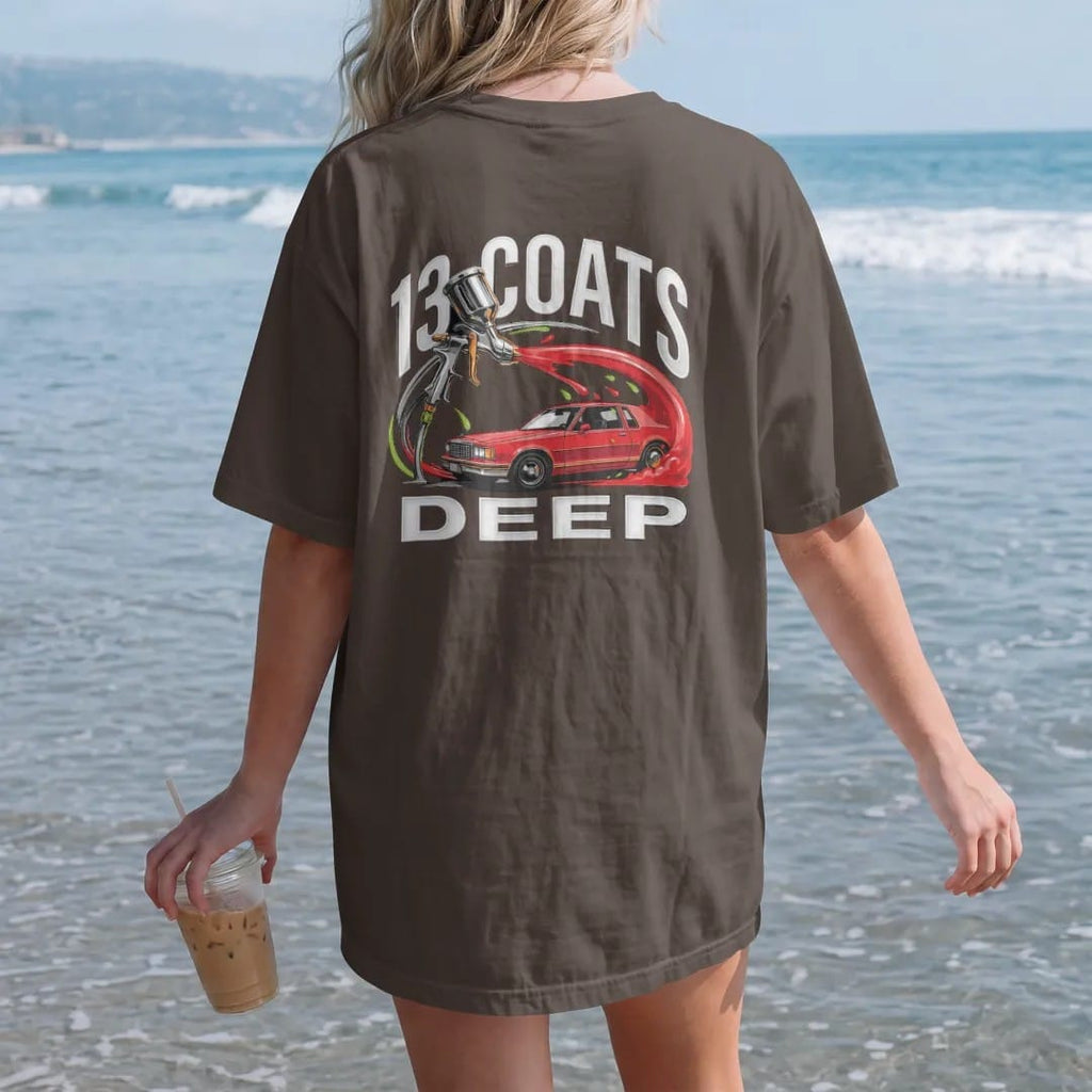 Woman on the beach in a Espresso tee, back view: a spray paint gun throwing an arc of candy paint that becomes the side of a boxy sedan, with 13 Coats over Deep in chrome letters.