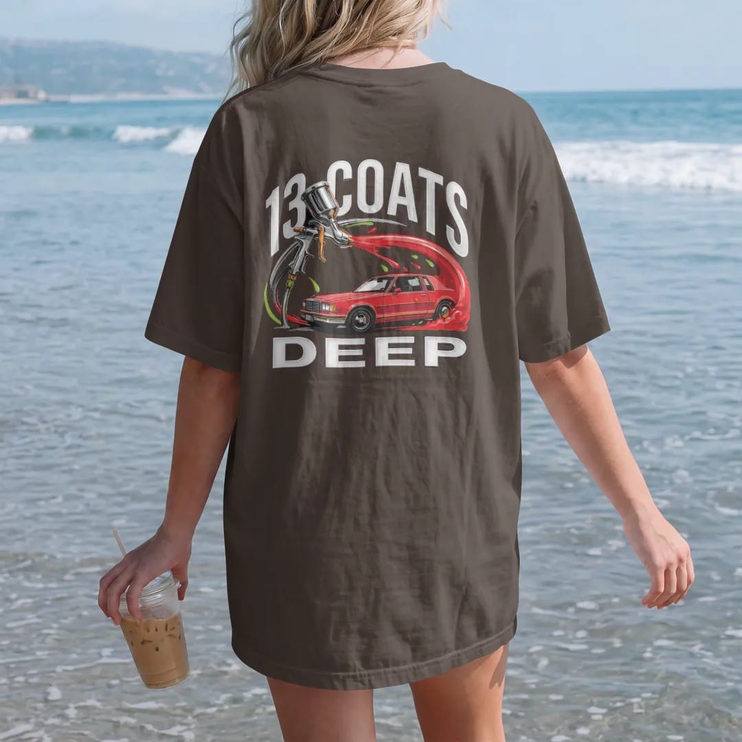 Woman on the beach in a Espresso tee, back view: a spray paint gun throwing an arc of candy paint that becomes the side of a boxy sedan, with 13 Coats over Deep in chrome letters.