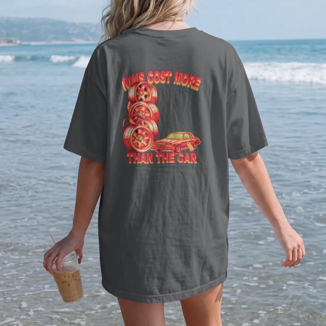Woman on the beach in a Graphite tee, back view: a tall stack of giant chrome rims next to a small faded boxy sedan with a price tag, under Rims Cost More and over Than The Car.