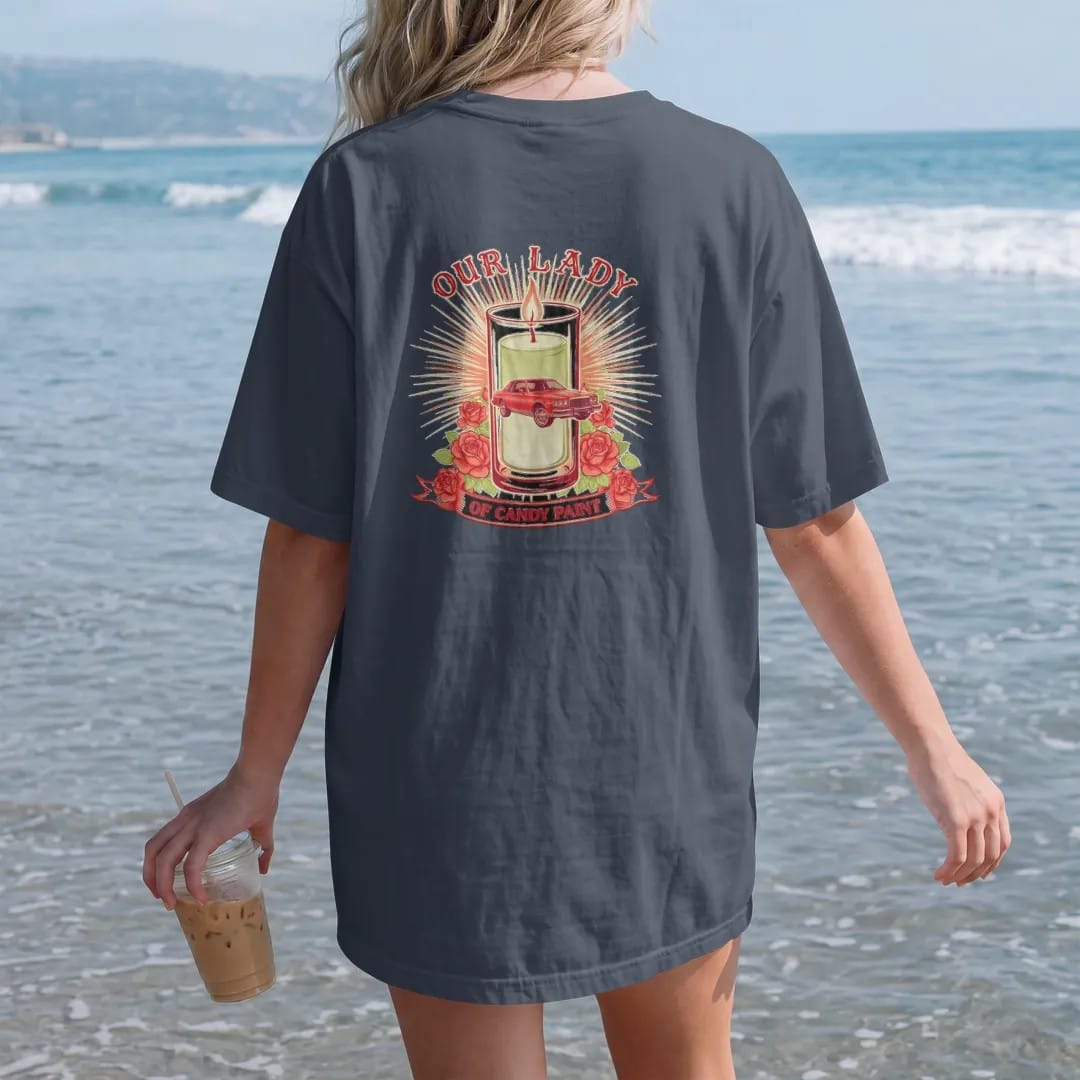 Woman on the beach in a Midnight tee, back view: a prayer-candle parody: a glowing votive with a boxy candy-red sedan on chrome wheels, roses and rays, Our Lady above and Of Candy Paint on a ribbon.