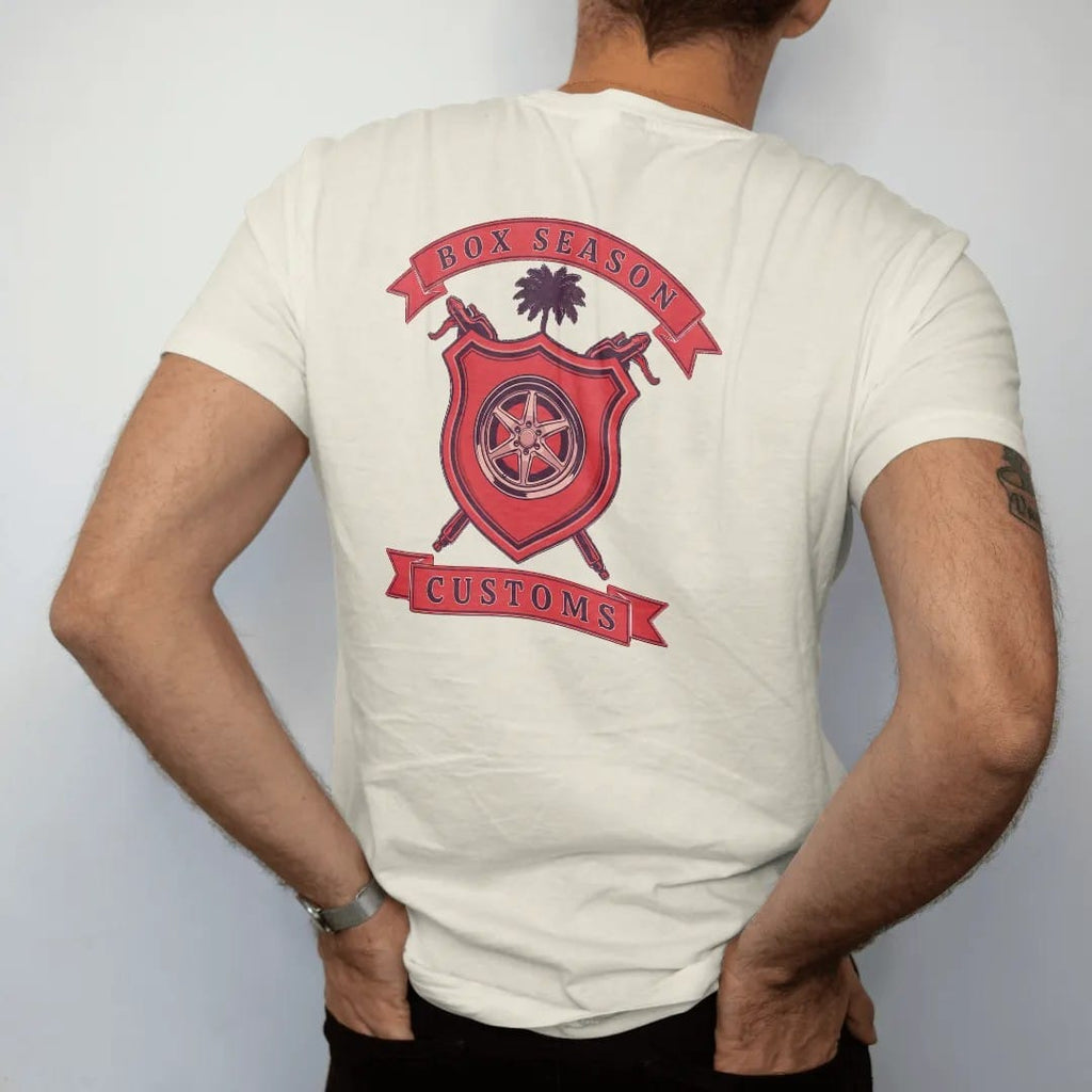 Man wearing a Ivory tee, seen from the back: a club crest: a red shield with a chrome wheel, crossed spray guns and a palmetto tree, Box Season and Customs on ribbon banners.