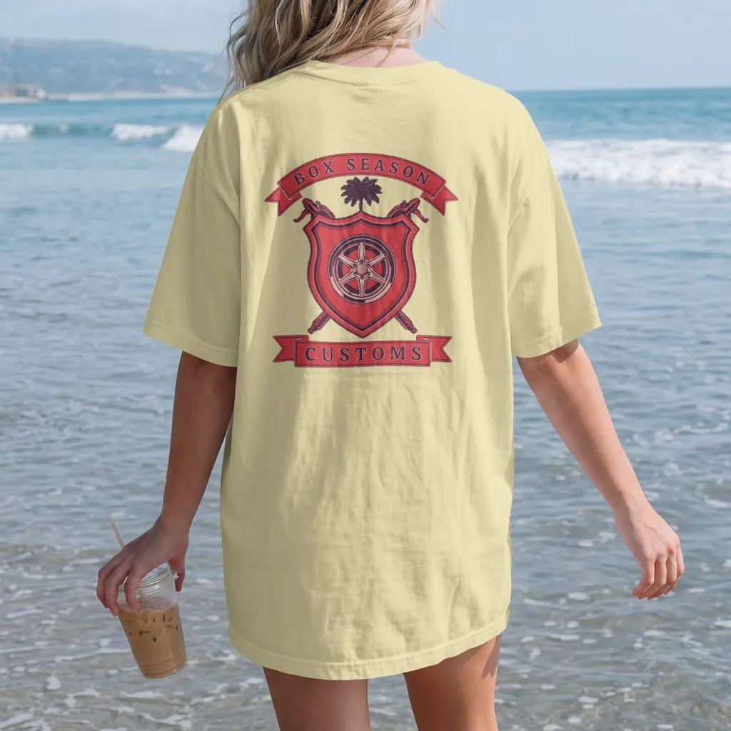 Woman on the beach in a Butter tee, back view: a club crest: a red shield with a chrome wheel, crossed spray guns and a palmetto tree, Box Season and Customs on ribbon banners.